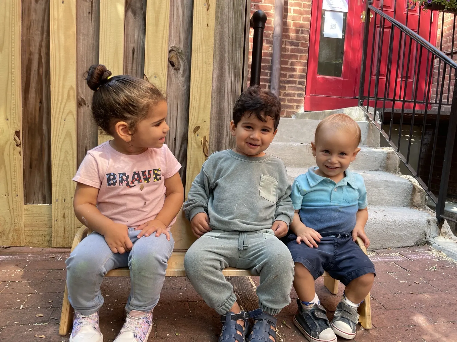 Three Starling toddlers sitting together on a bench at the school's red front door on Capitol Hill